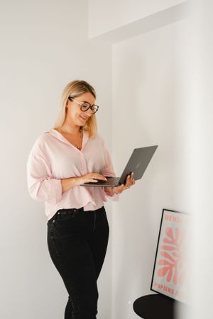 Person in pink blouse and glasses working on laptop against white wall with minimal decor.