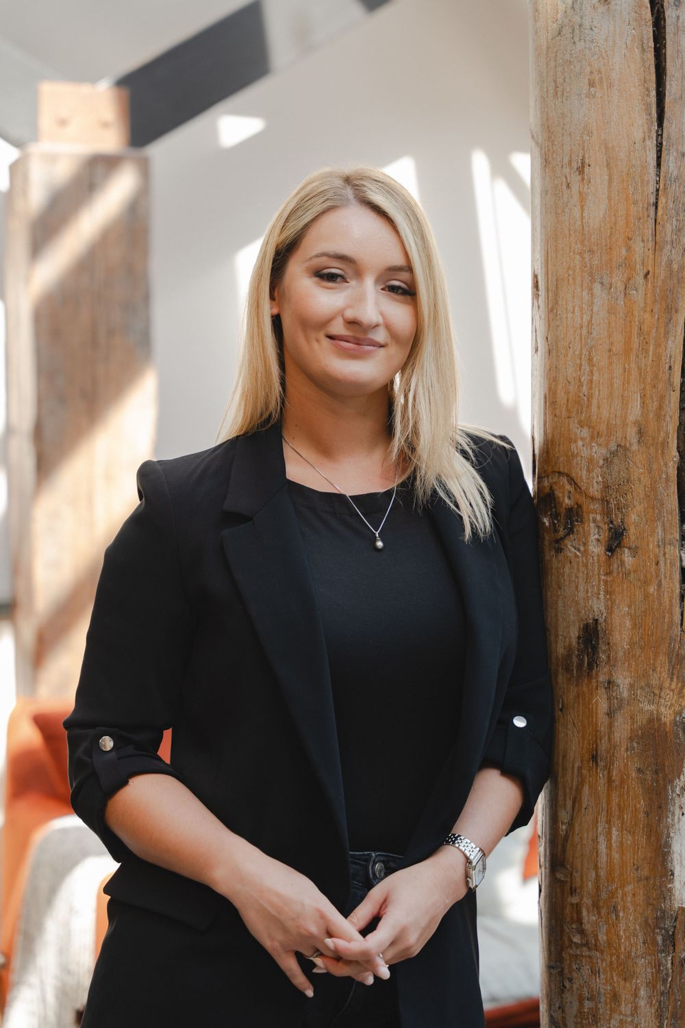 Professional portrait in black blazer standing by wooden beam with natural lighting.