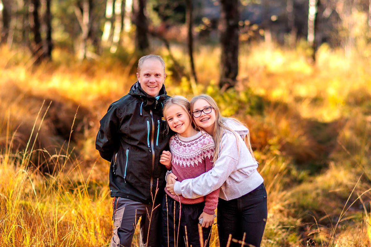 Far og to døtre står sammen i en solfylt skog i høstfarger. Storesøster holder rundt lillesøster.