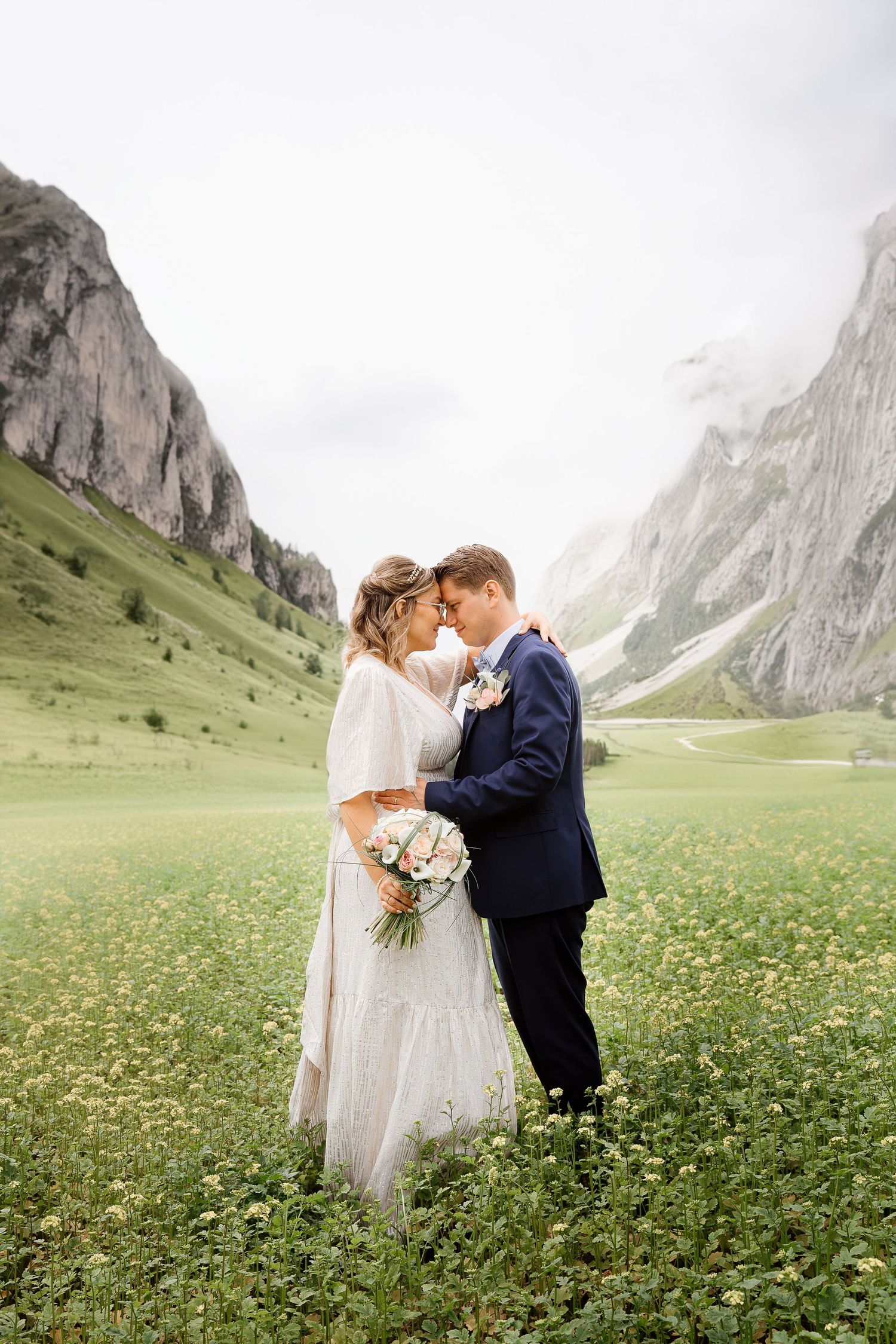 Wedding couple embracing in scenic mountain meadow with dramatic peaks in background.