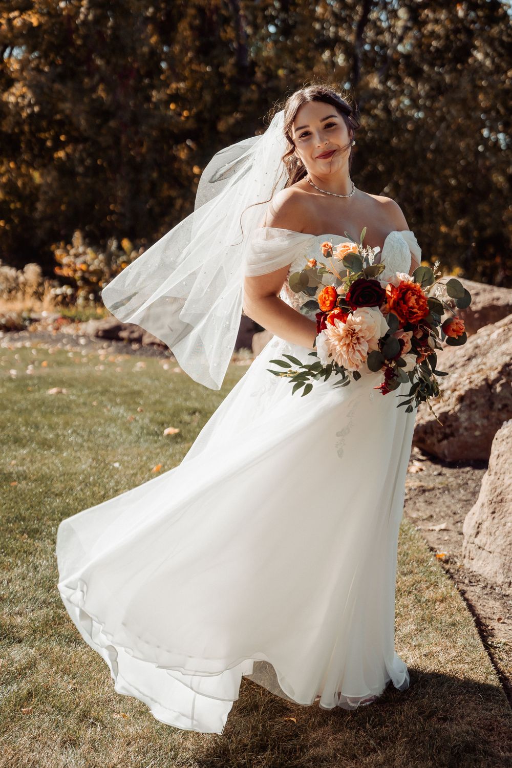 A bride in a flowing white off-shoulder gown holds a vibrant autumn bouquet against a rustic outdoor backdrop.