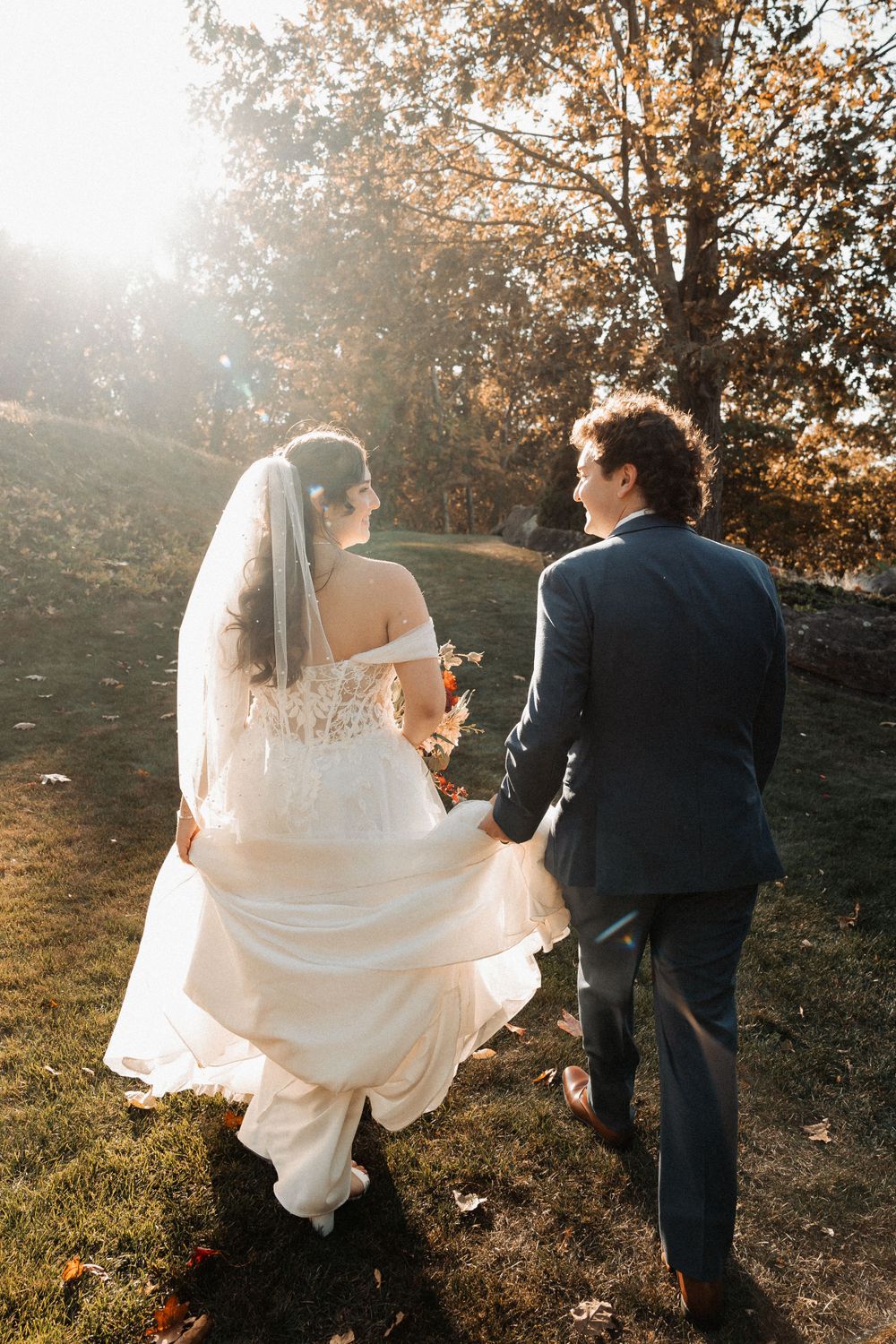 Couple walking hand in hand during golden hour with trailing wedding dress and autumn trees.