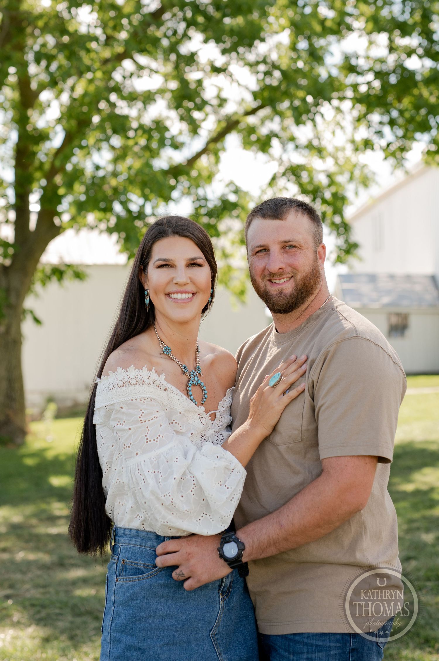 Couple poses outdoors in casual attire against green trees and white barn background.