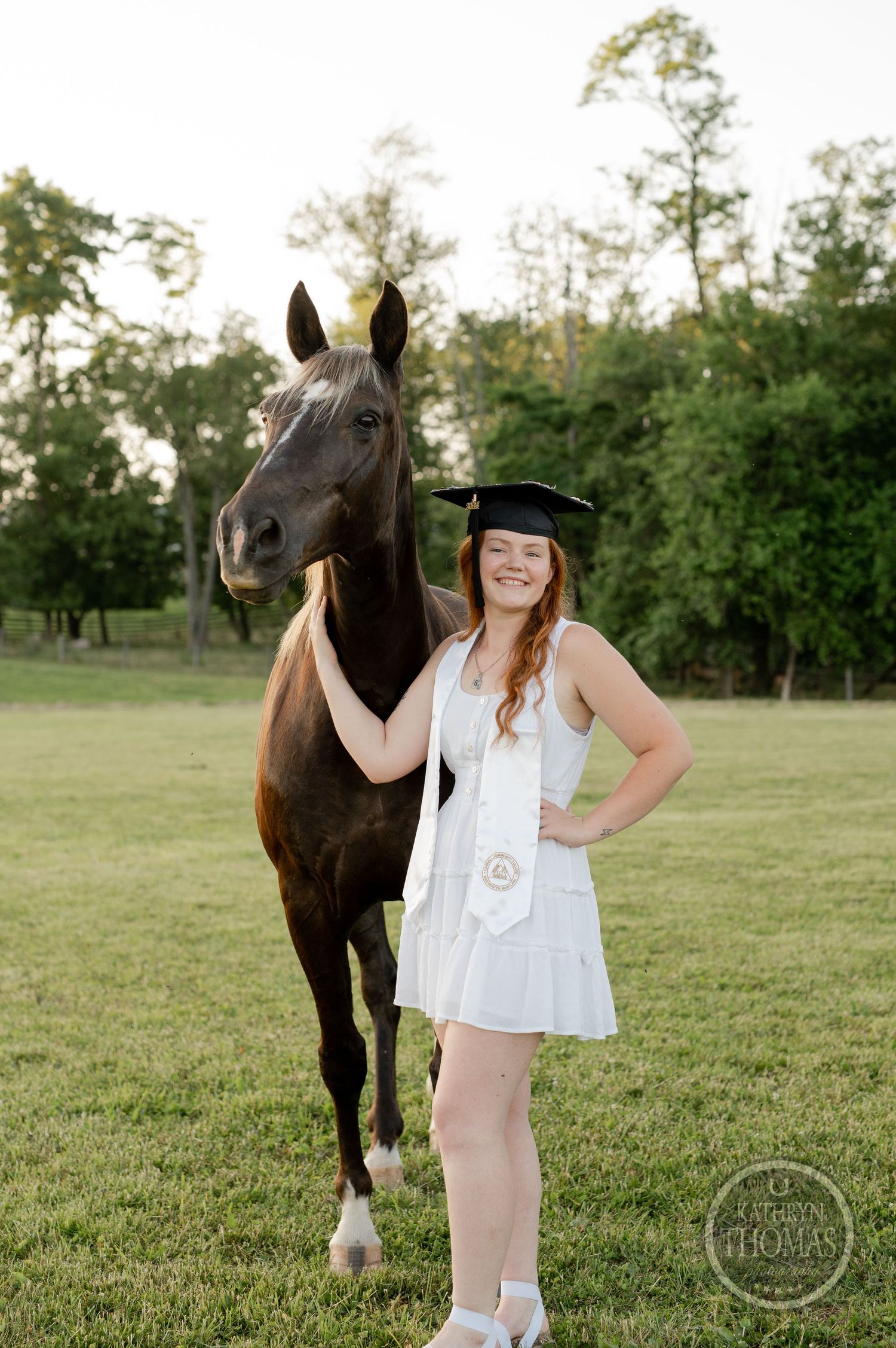 Graduate in white dress stands beside dark horse in pastoral setting.