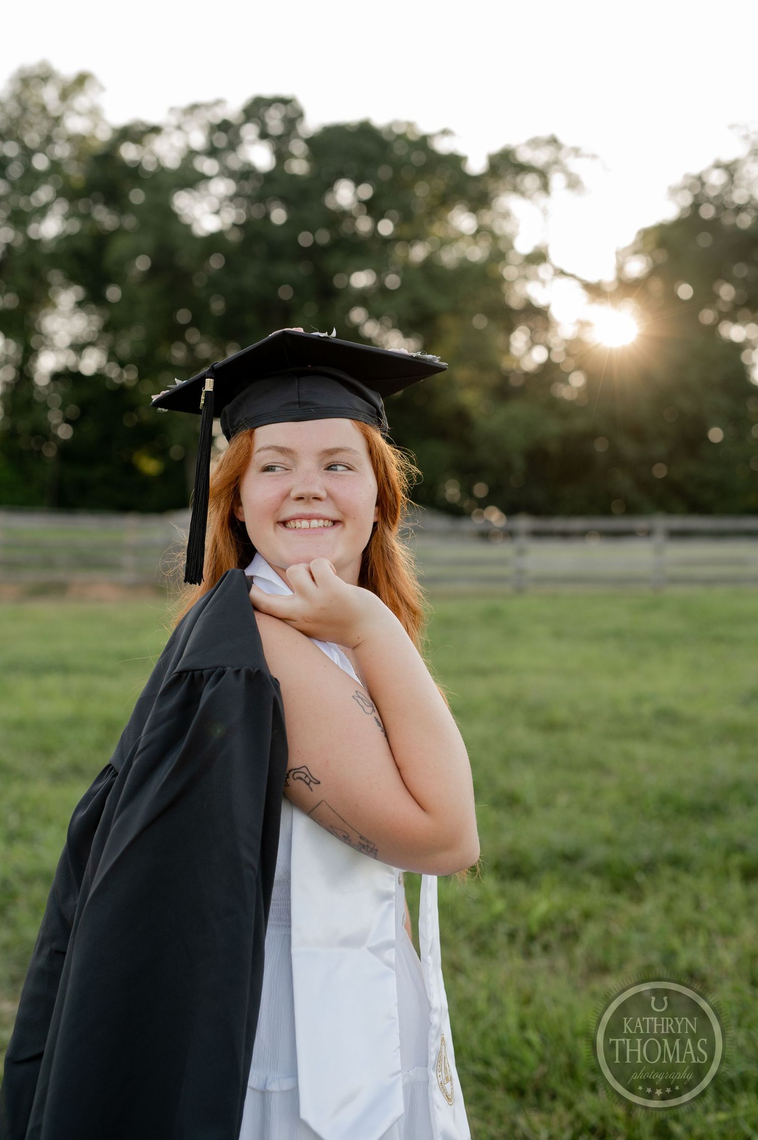 Graduate in traditional academic regalia poses against sunset in pastoral setting.