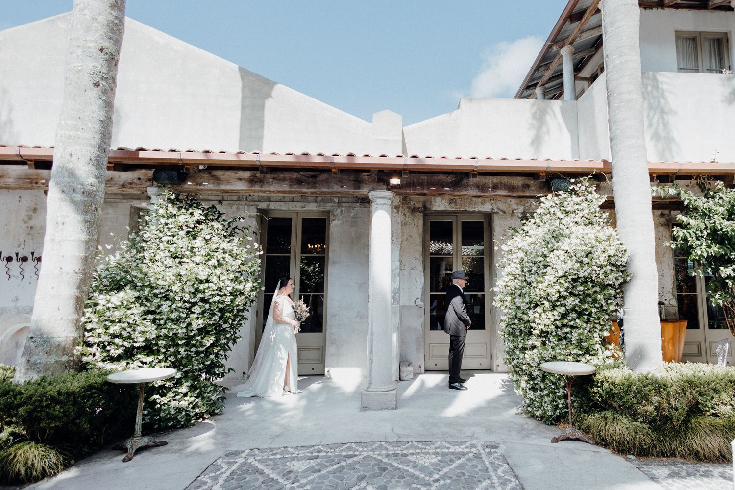 First Look of a bride and her father in a courtyard with blooming shrubs. Captured by Apertured Media Auckland Wedding Photographer