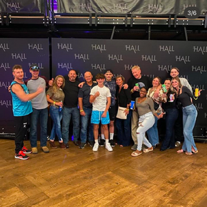 A group of people posing together against a dark wall with repeating HALL text in a social gathering setting.