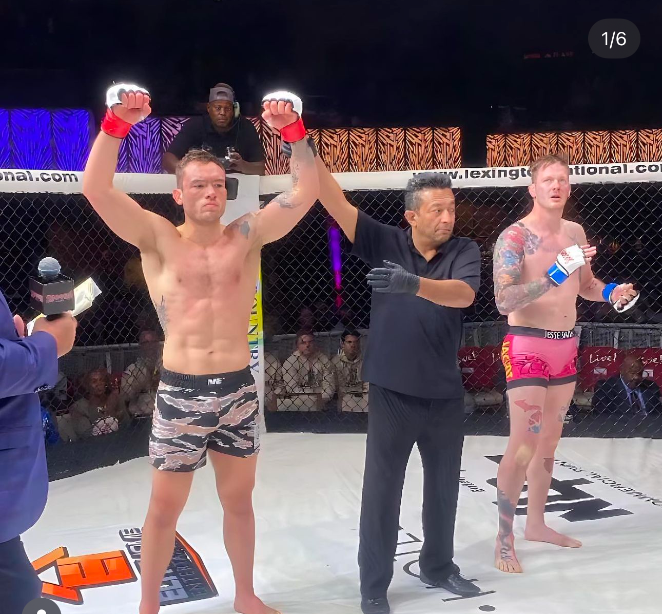 A victorious MMA fighter raises their arms while wearing camo shorts in an octagon cage during a competition match.