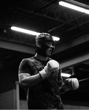 A boxer in protective headgear and gloves stands ready in black and white under fluorescent gym lights.