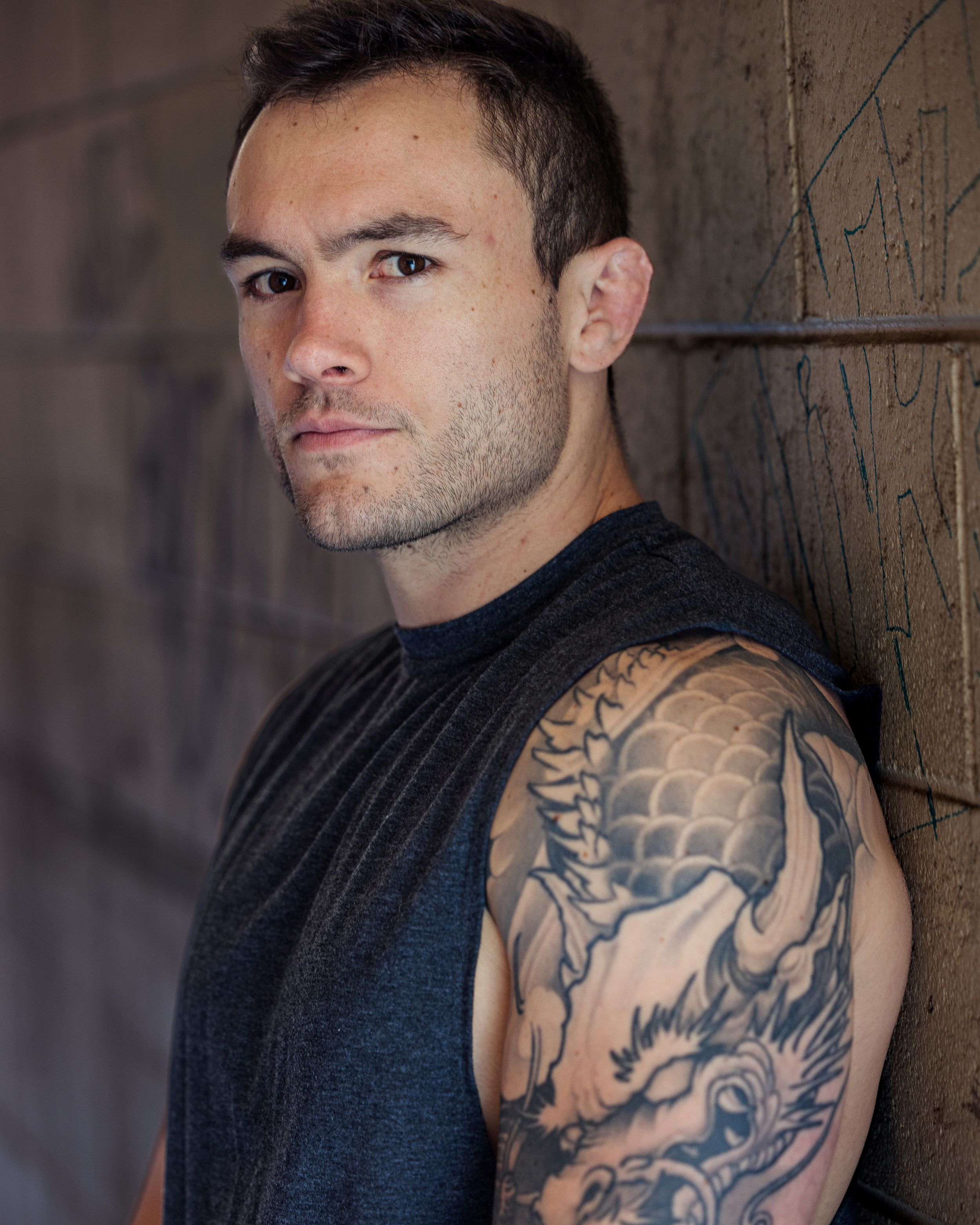 Athletic person in black sleeveless top shows detailed dragon tattoo sleeve while posing against textured wall.