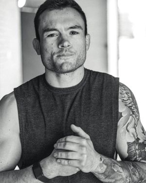Black and white portrait of a muscular man in a sleeveless shirt with visible tattoos on his arms.