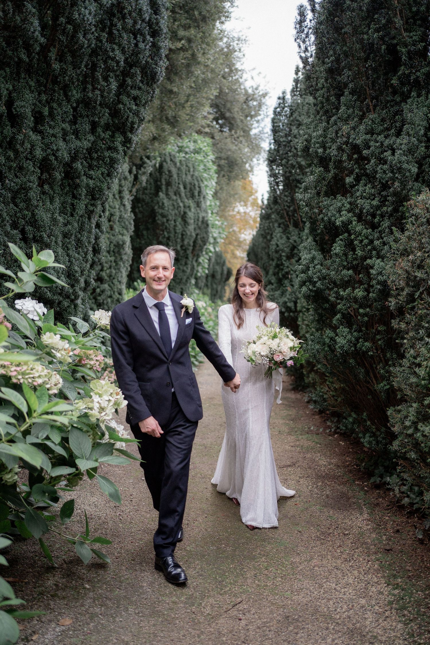 A wedding couple walks hand in hand down a garden path lined with tall cypress trees. Hotel Endsleigh, Devon wedding photography.
