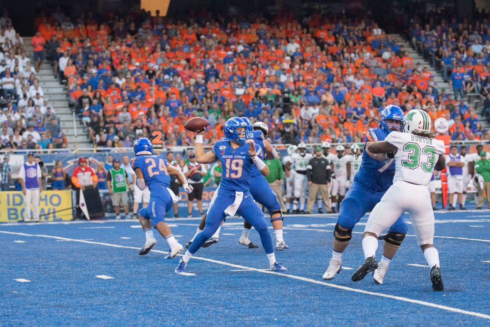 College football players in blue and white uniforms compete on a blue field with orange-clad fans in stadium stands.
