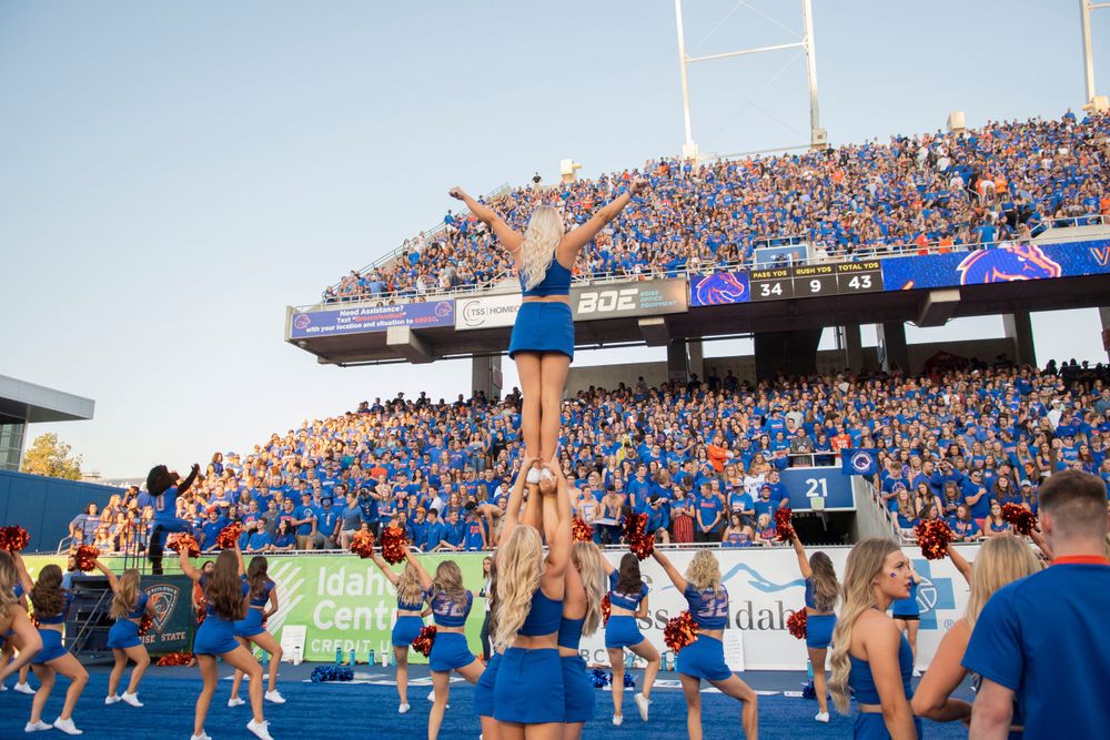 Cheerleaders perform a high-flying stunt in front of crowded stadium bleachers filled with fans wearing blue.