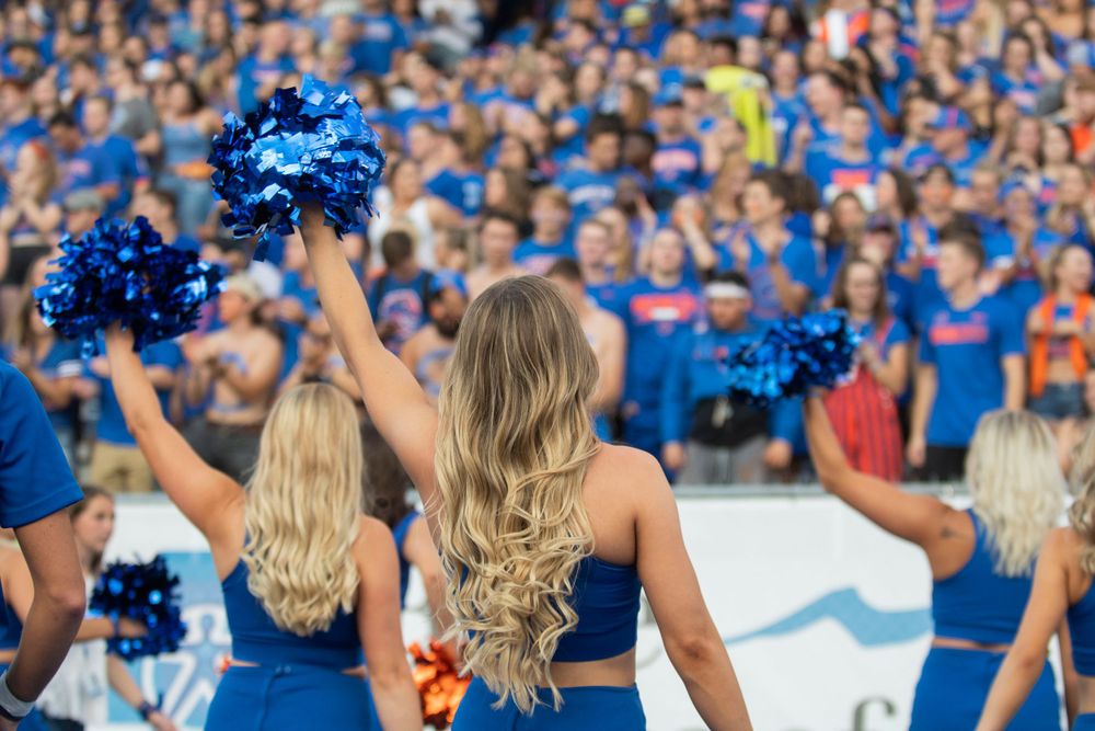 College cheerleaders in blue uniforms wave pom-poms in front of a large crowd of fans at a sporting event.