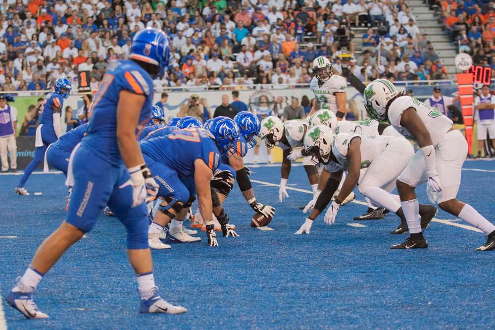 Football teams line up at scrimmage on blue turf field with offensive and defensive players facing off.
