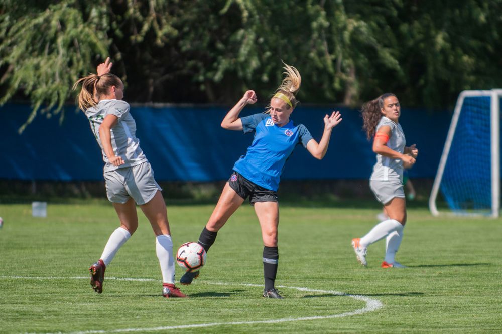Women soccer players compete for possession of the ball during an intense match on a sunny grass field.