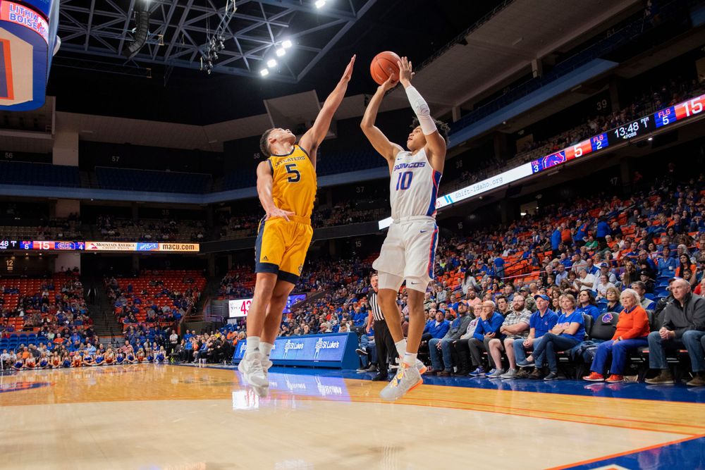 Two basketball players jump during a game, with one attempting to block the other's shot at a college arena.