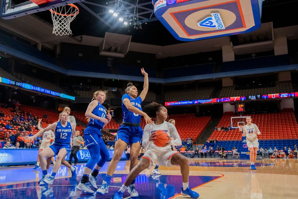 Basketball players compete for a rebound during an intense game in an indoor arena with bright lighting and empty seats.