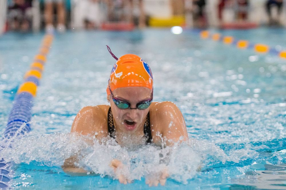 Competitive swimmer performs breaststroke in swimming pool with orange swim cap and goggles during race.