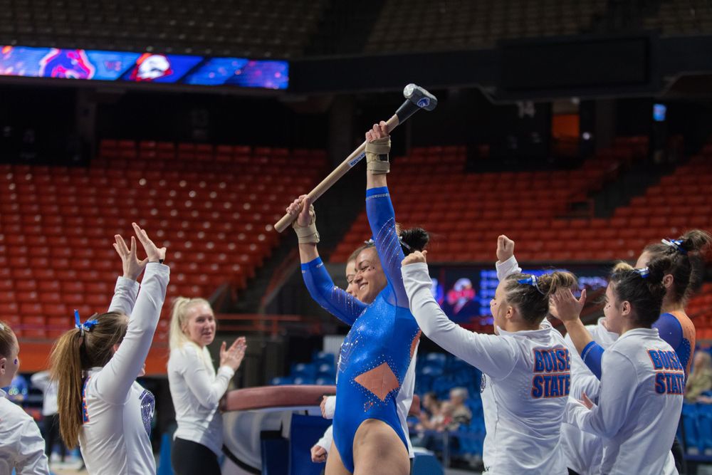 Gymnast in blue leotard celebrates with teammates holding up high scores at competitive event in arena.
