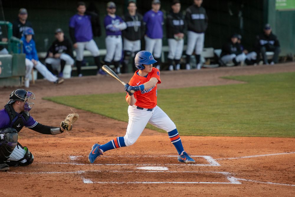 Baseball batter in orange and blue uniform swings at pitch during nighttime game with catcher and dugout in background.