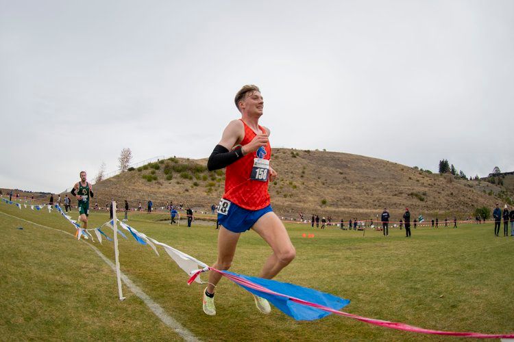 Runner in orange and blue uniform competes on grassy cross country course with hills in background.