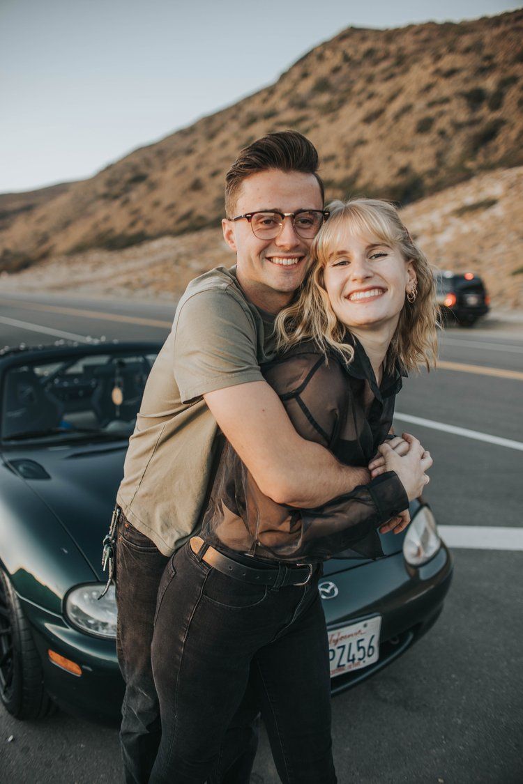 A happy couple embraces on a desert highway next to a black Mazda Miata convertible during a sunny day.