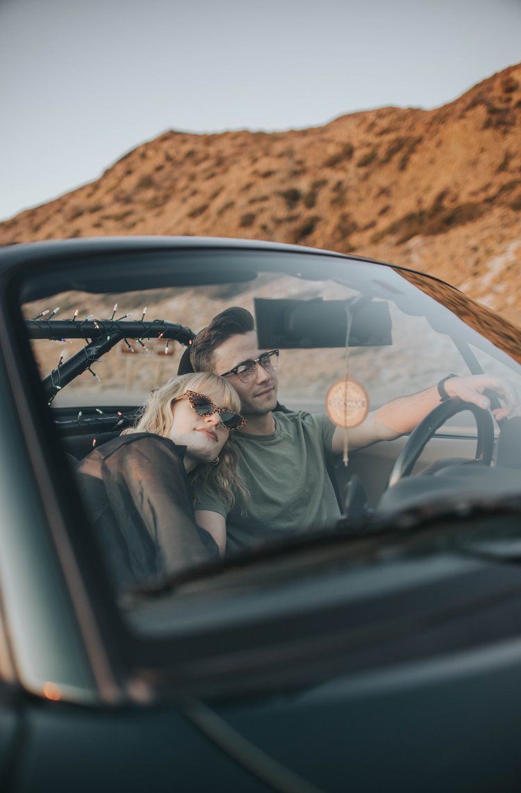 A couple relaxes in a parked car against a mountainous desert backdrop during golden hour.
