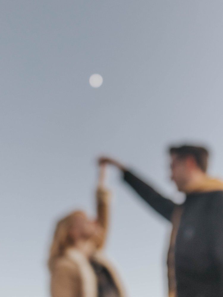 Two blurred figures reach upward toward a glowing full moon in the misty gray sky.