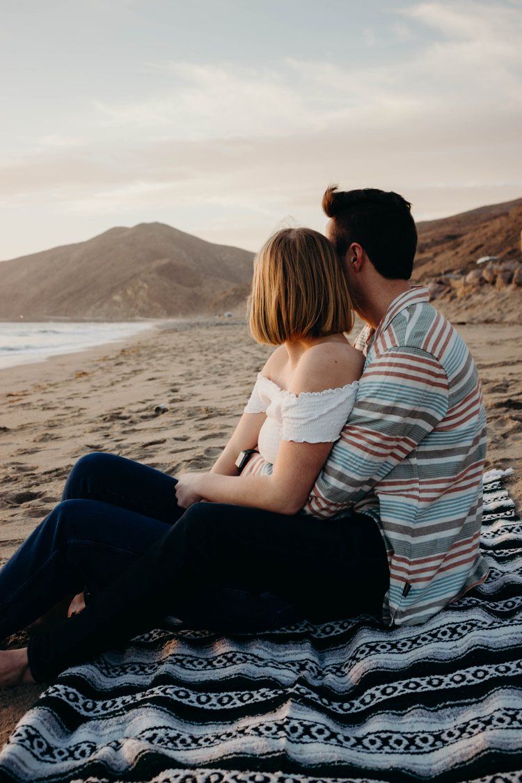 A couple sits together on a patterned blanket at the beach during sunset, overlooking mountains and waves.