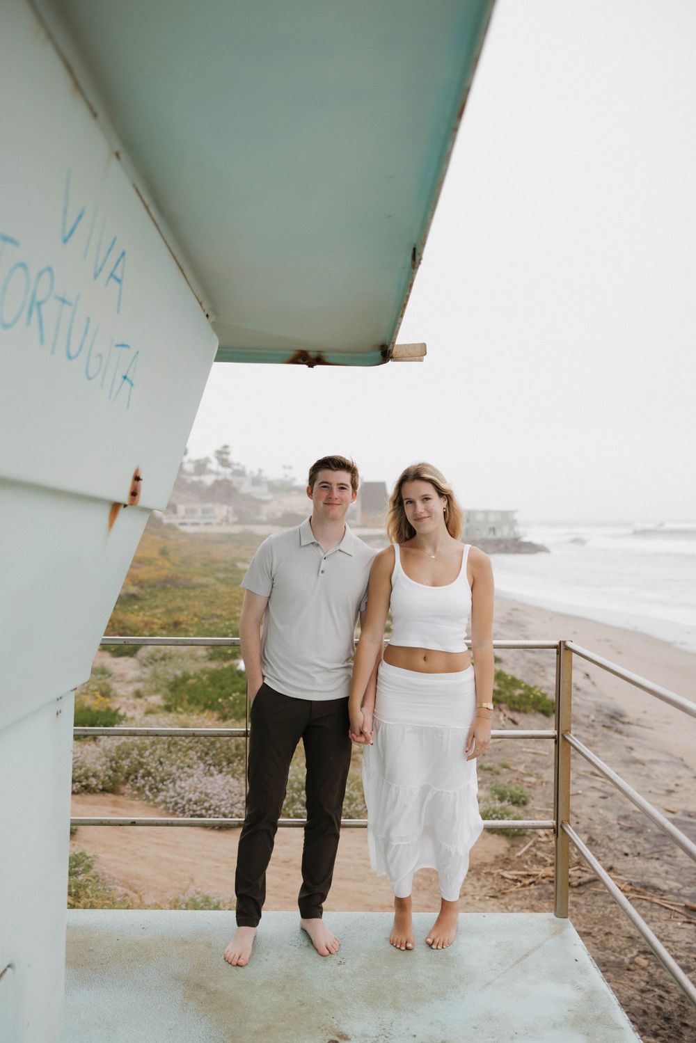 A couple in casual white outfits stand barefoot on a beach overlook area near the ocean on an overcast day.