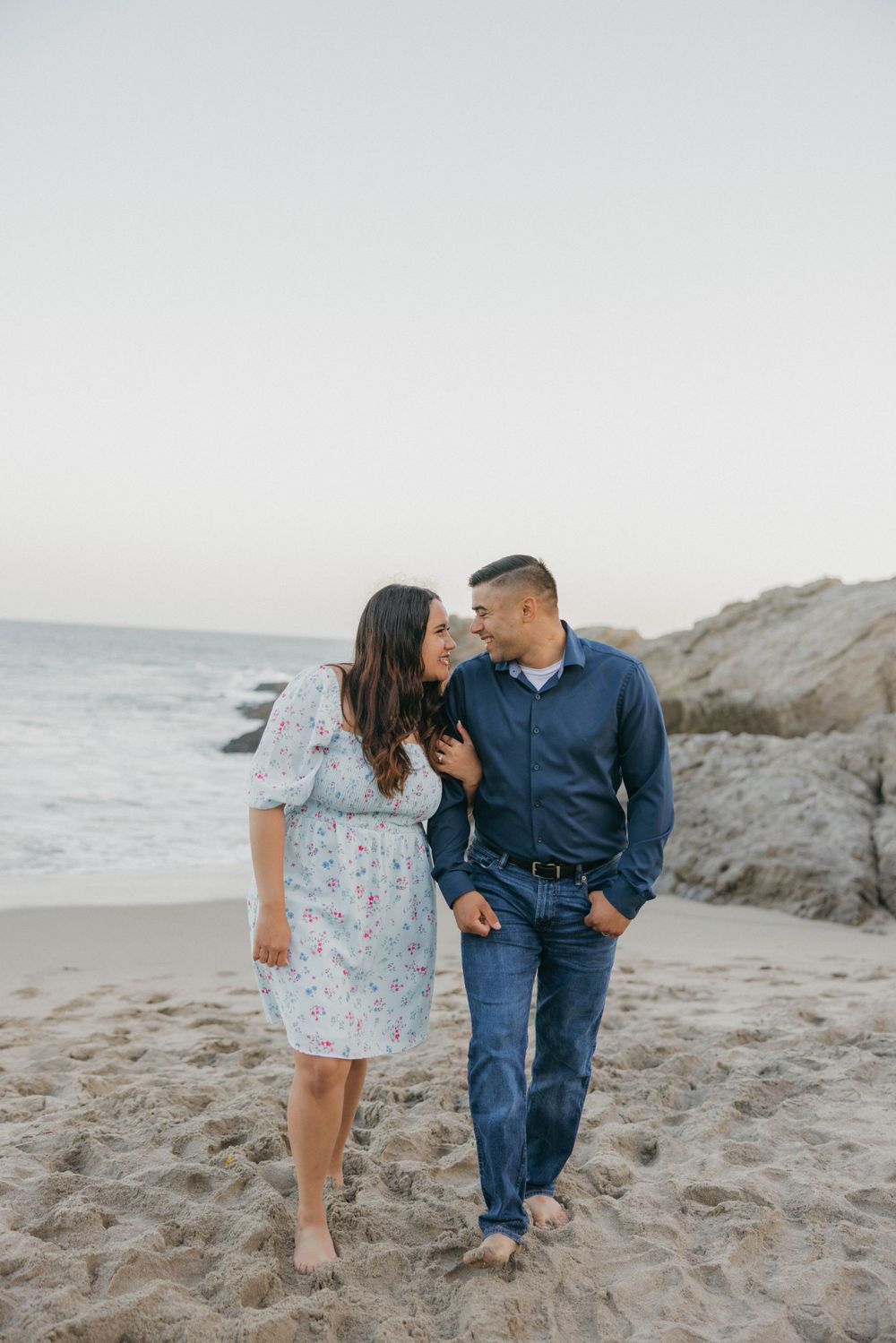 A romantic couple walks together on a sandy beach at sunset with ocean waves and rocky cliffs in the background.