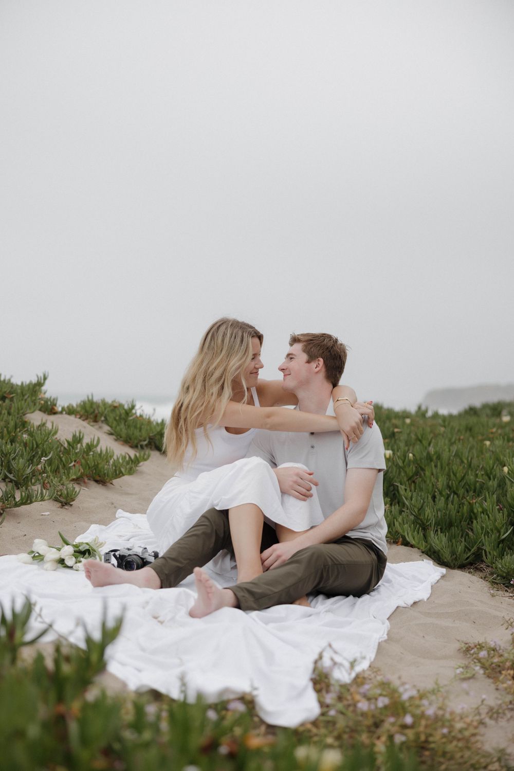 A romantic couple sharing an intimate moment while sitting on a white blanket surrounded by coastal vegetation.