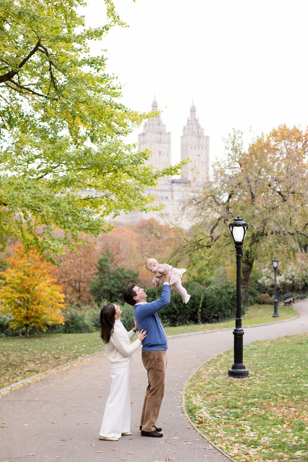 A couple dances playfully on a path in Central Park during autumn with fall foliage and historic buildings in background.