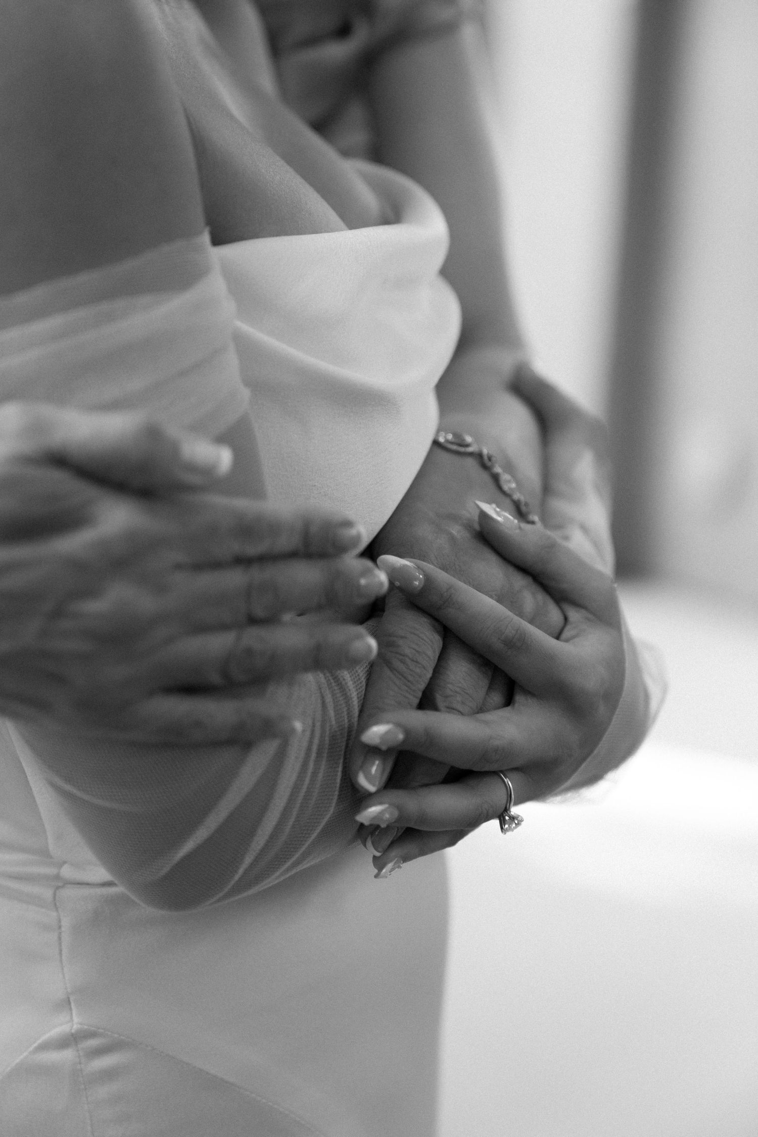Black and white photo showing hands clasped together over a wedding dress with delicate beading and lace details.