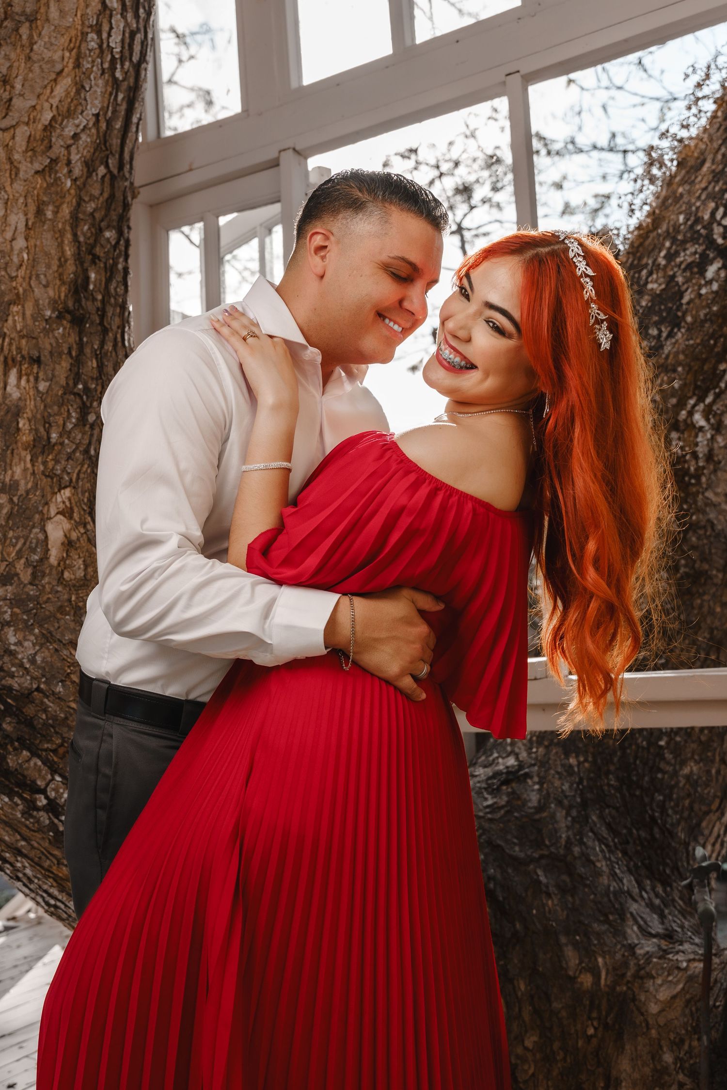 A romantic couple embraces outdoors, with her wearing a stunning off-shoulder red pleated dress and him in a white dress shirt.