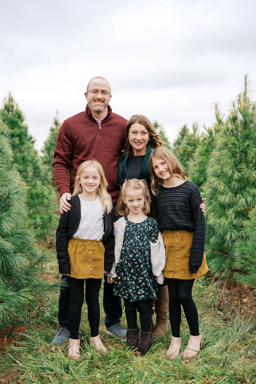 Family poses together among rows of evergreen trees at Christmas tree farm.