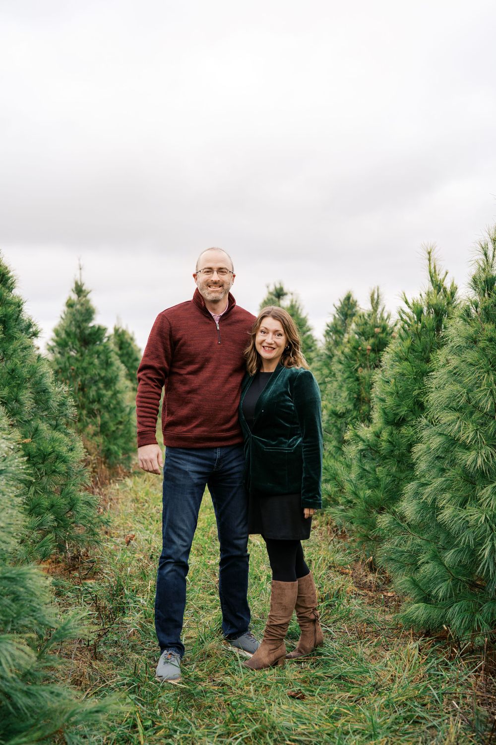 A couple stands together in a Christmas tree farm wearing burgundy and dark green.