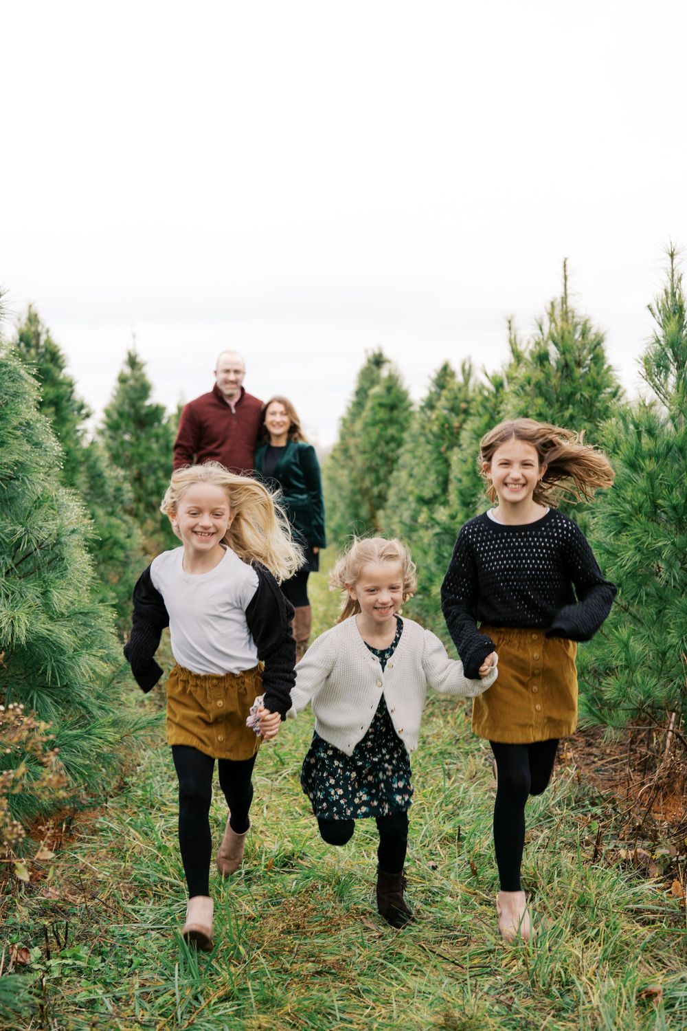 Three children run joyfully through a Christmas tree farm while adults watch in the background.