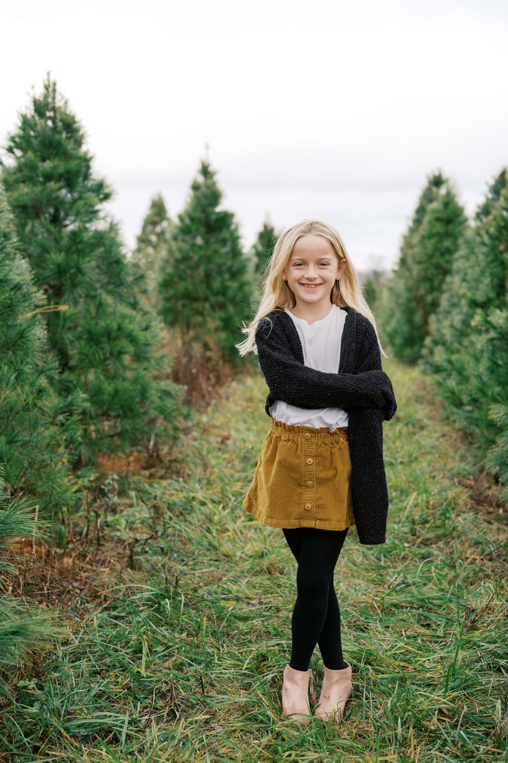 A young person in a mustard skirt and black cardigan poses confidently among pine trees.