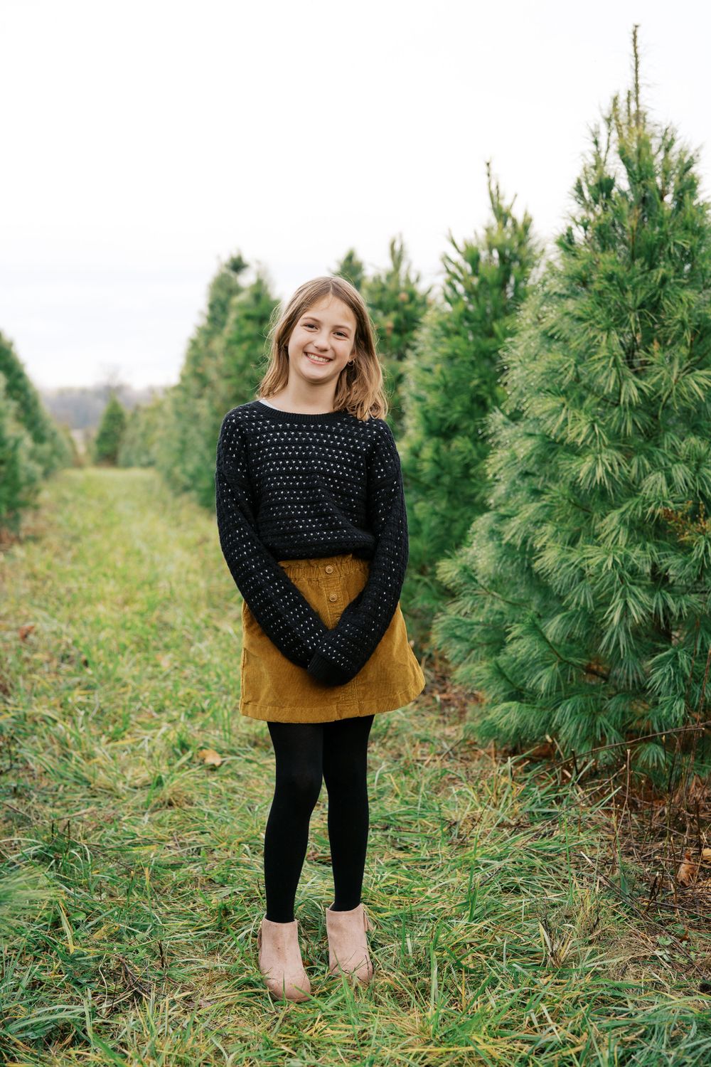 A person in a black sweater and mustard skirt stands between rows of evergreen trees.