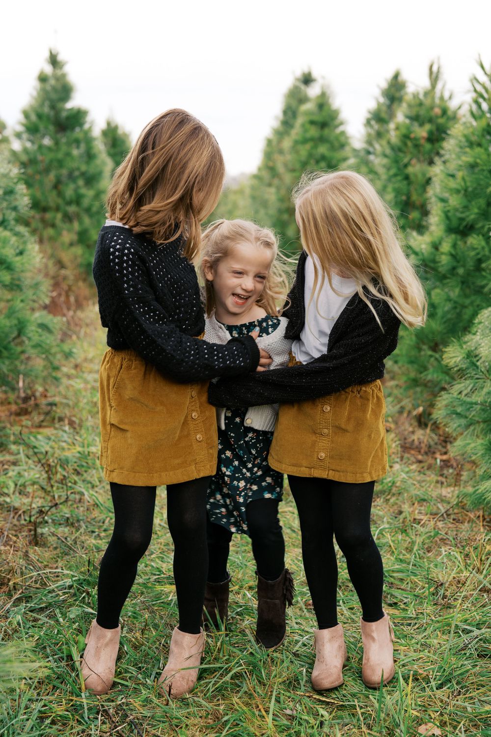 Three people in matching mustard skirts share a candid laughing moment among pine trees.