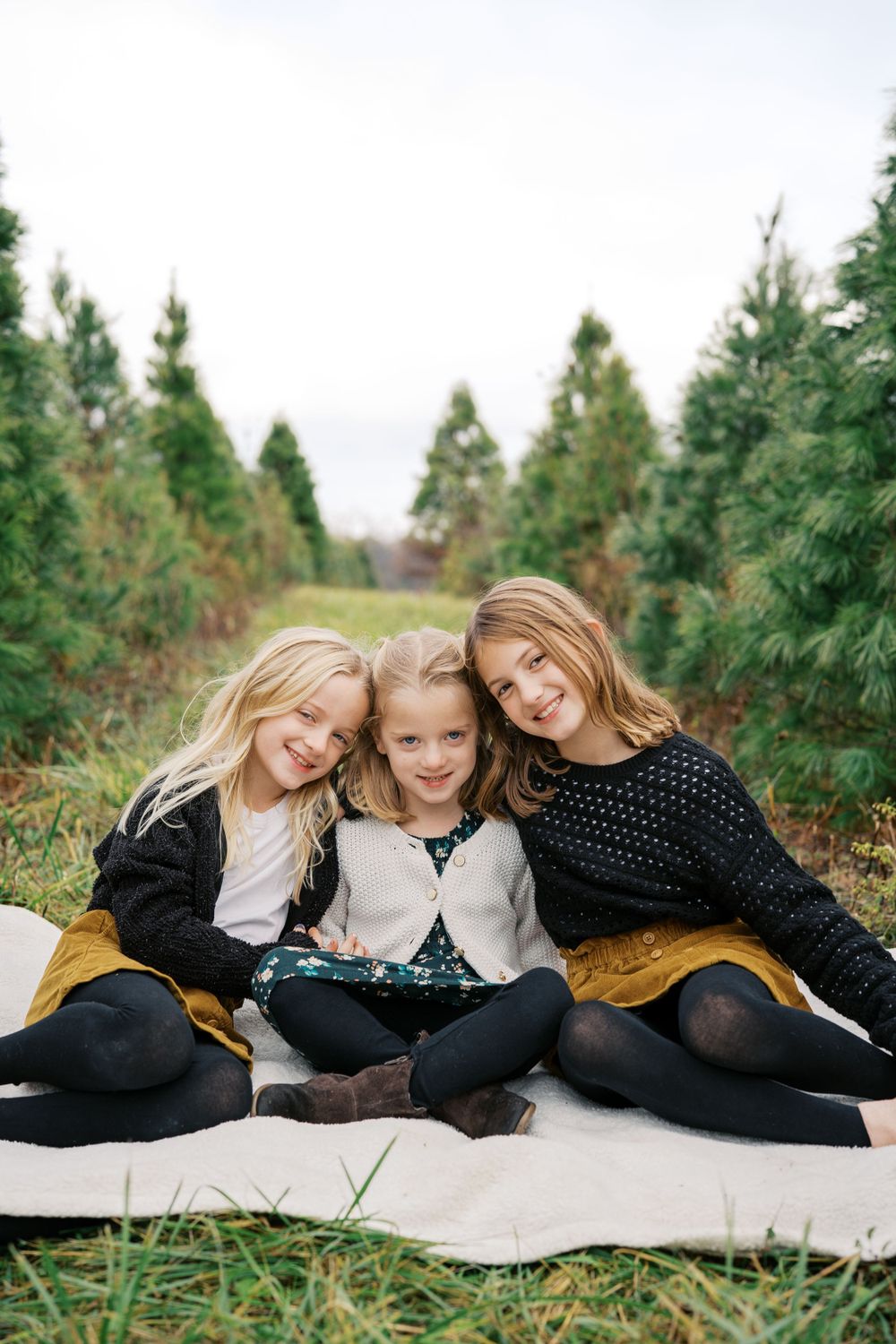 Three children in winter outfits lying on a white blanket among evergreen trees.