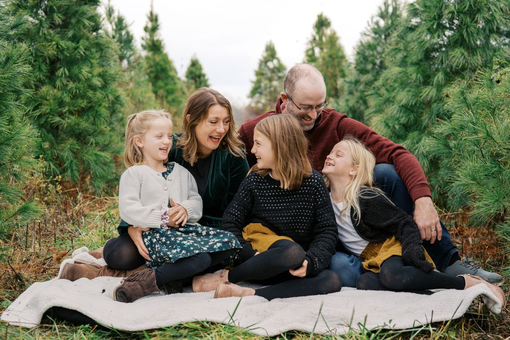 Three children in winter outfits lying on a white blanket among evergreen trees.