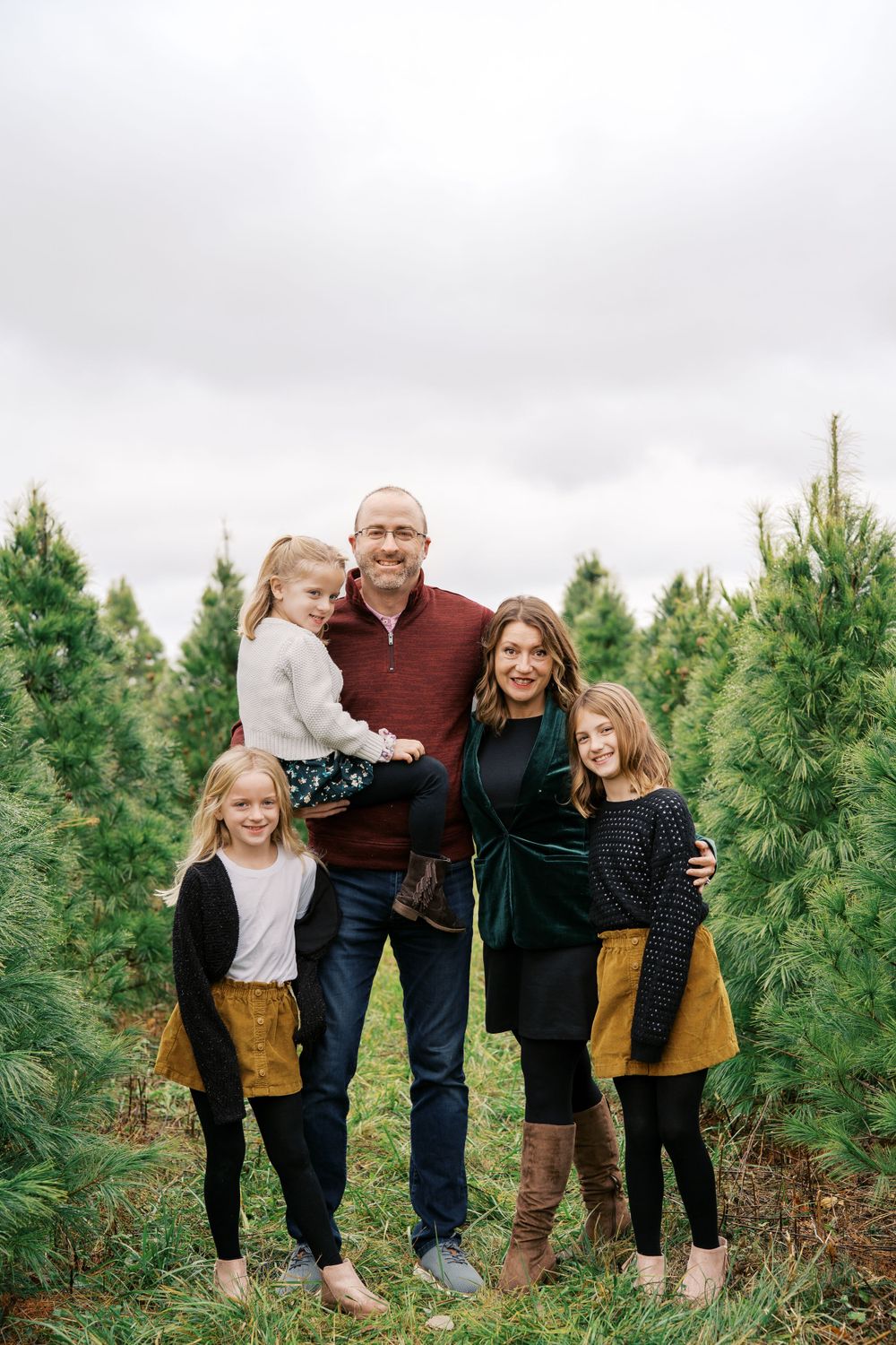 A family poses together among evergreen trees wearing coordinated fall outfits in burgundy, black and mustard yellow.