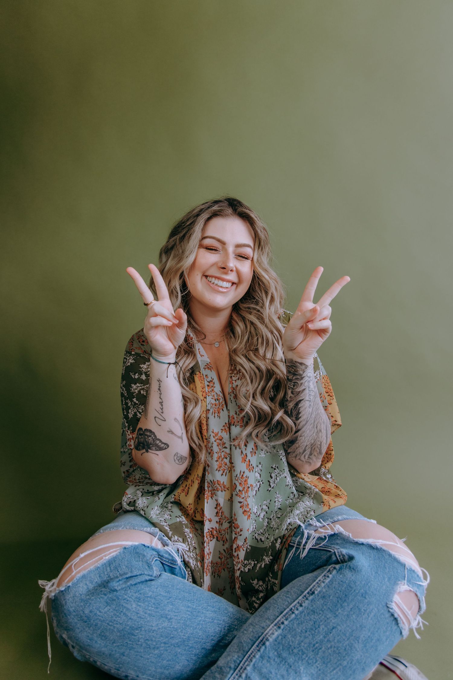 Person in patterned top and ripped jeans making peace signs while sitting casually.