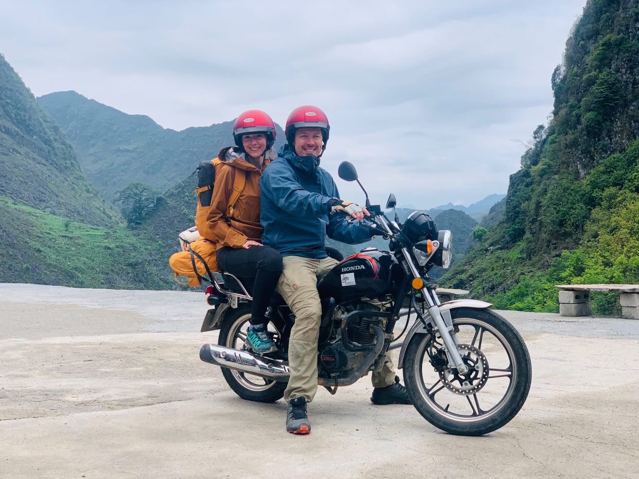 Evan and Kristen riding a motorcycle along a scenic mountain road with green peaks and cliffs in the background in northern Vietnam