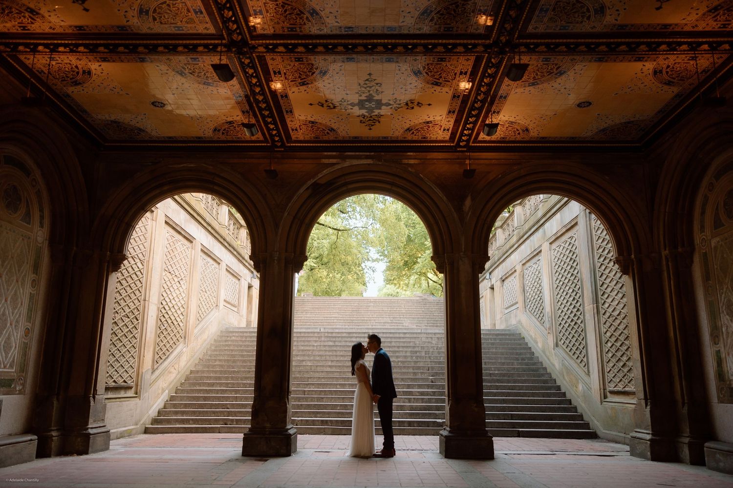 A romantic wide shot of a couple facing each other beneath a bridge, framed by dramatic architecture and soft natural light.