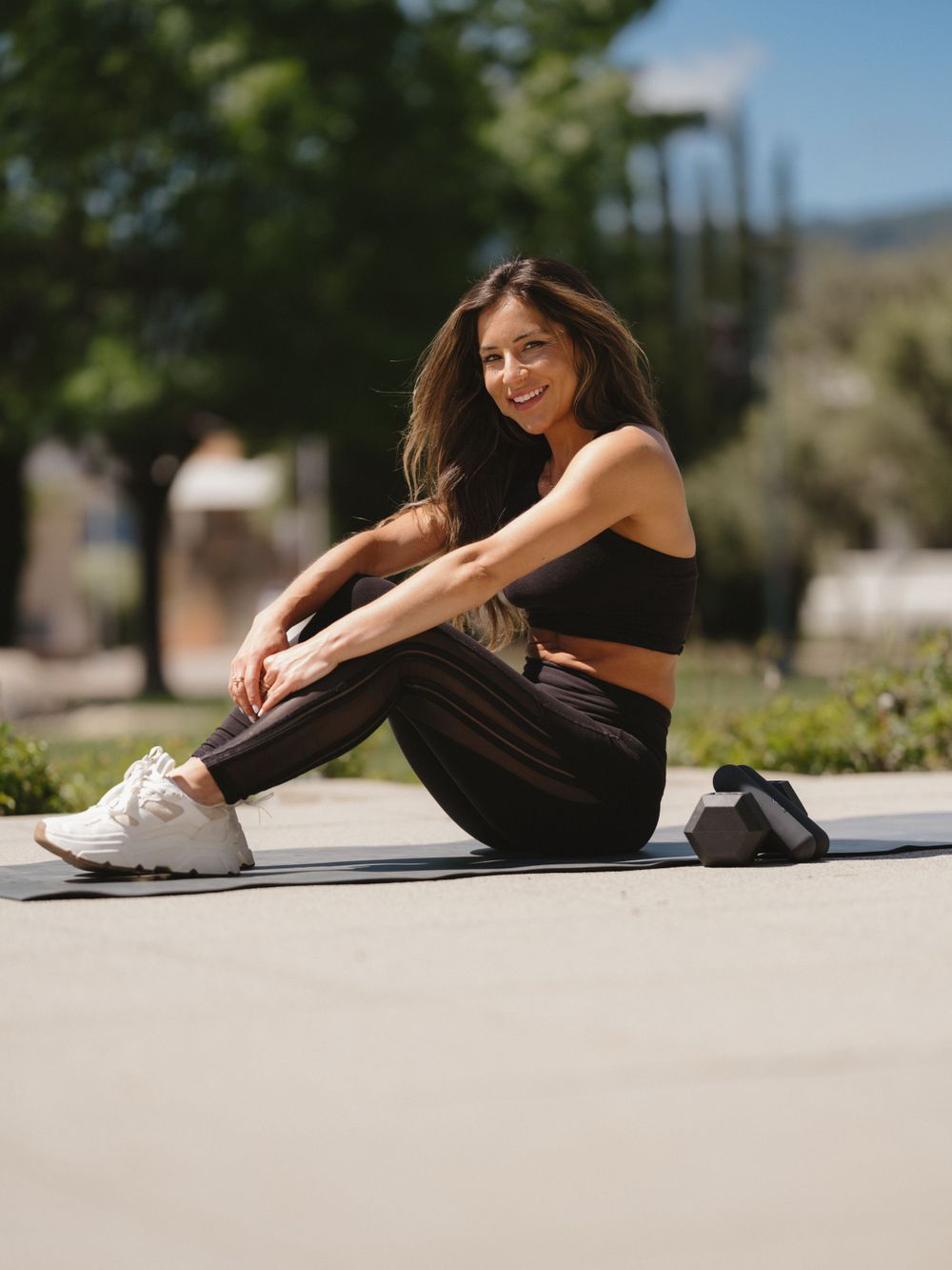 Fitness model in black workout clothes stretches on yoga mat in sunny outdoor setting.